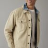 Man wearing a tan overshirt coat with a plaid collar over a grey sweater.